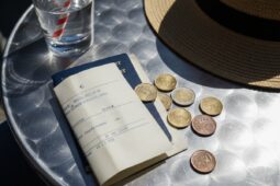 Glas Wasser, Reisepass, Rechnung und Euro-Münzen auf einem Metalltisch neben einem Strohhut